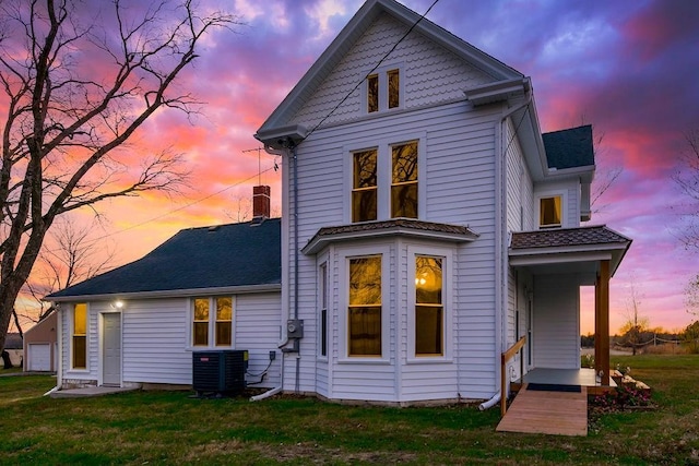 rear view of property featuring a shingled roof, central AC unit, a chimney, a garage, and a yard