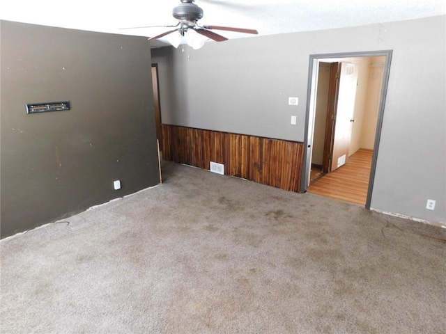 carpeted spare room featuring wooden walls, a ceiling fan, visible vents, and wainscoting