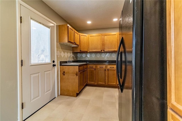 kitchen featuring brown cabinetry, freestanding refrigerator, a sink, decorative backsplash, and dark countertops