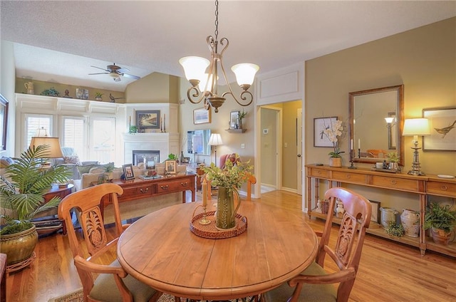 dining space with ceiling fan with notable chandelier, a fireplace, light wood-style floors, and vaulted ceiling