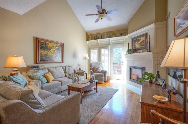 living room featuring high vaulted ceiling, a fireplace, ceiling fan, a textured ceiling, and light wood-type flooring