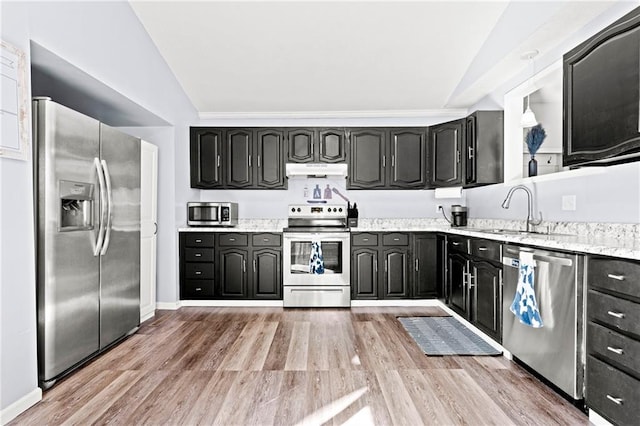 kitchen with a sink, under cabinet range hood, vaulted ceiling, stainless steel appliances, and dark cabinets