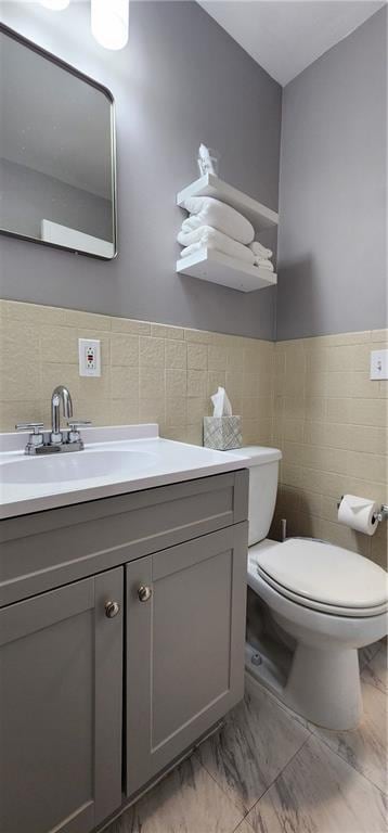 half bath with a wainscoted wall, toilet, marble finish floor, tile walls, and vanity