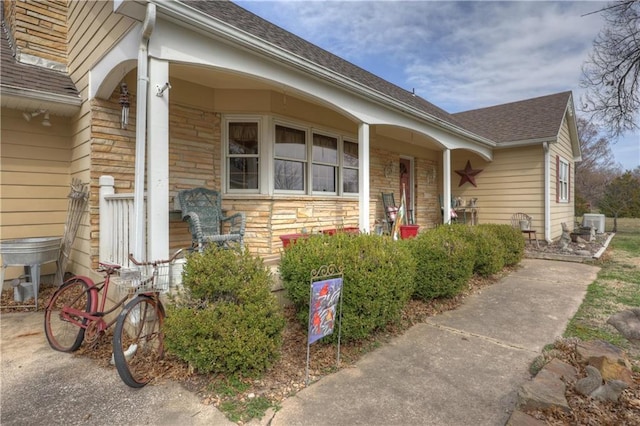 doorway to property featuring cooling unit, stone siding, roof with shingles, and covered porch