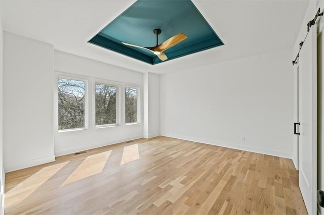 empty room featuring a raised ceiling, a ceiling fan, a barn door, light wood-style floors, and baseboards