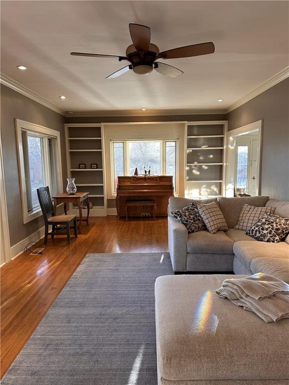 living room featuring built in shelves, ornamental molding, a ceiling fan, wood finished floors, and baseboards