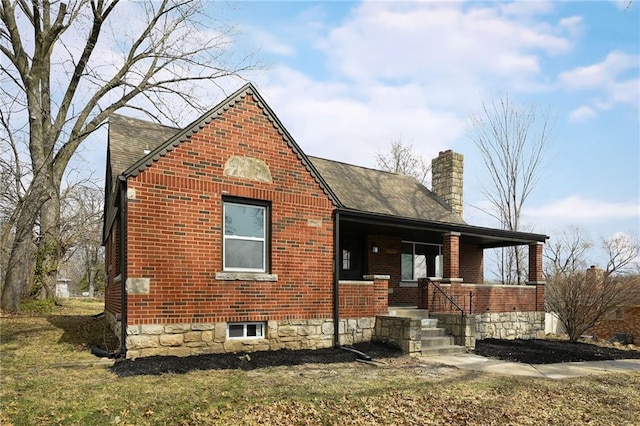 view of front facade featuring brick siding, covered porch, a chimney, and roof with shingles