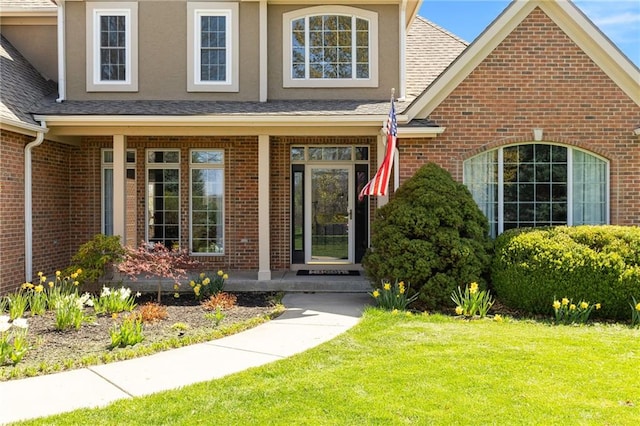entrance to property featuring a shingled roof, brick siding, stucco siding, and a yard
