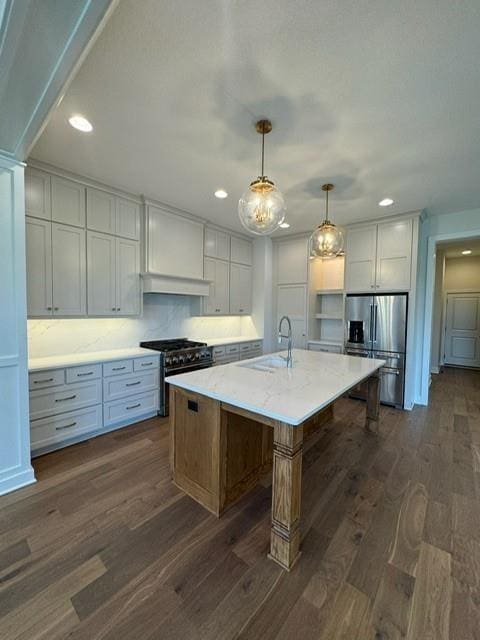 kitchen with an island with sink, a sink, dark wood-type flooring, appliances with stainless steel finishes, and pendant lighting