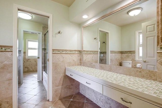 full bath featuring a shower with door, wainscoting, tile walls, toilet, and tile patterned floors