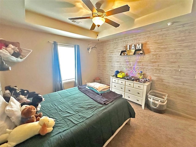 carpeted bedroom featuring a tray ceiling, wooden walls, visible vents, and ceiling fan