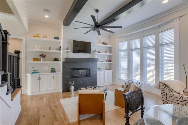 living room with built in shelves, a ceiling fan, ornamental molding, a tile fireplace, and light wood-style floors