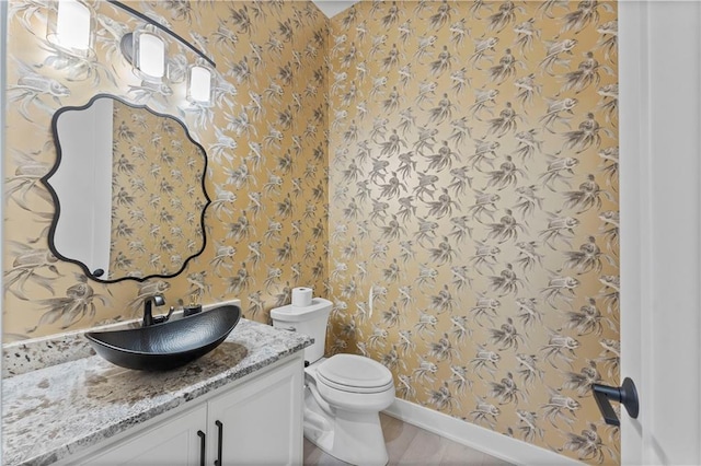 half bathroom with baseboards, toilet, vanity, and wallpapered walls