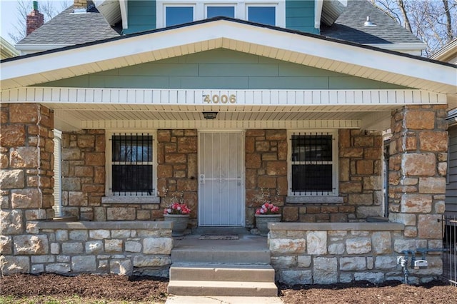 doorway to property with stone siding, a porch, and a shingled roof