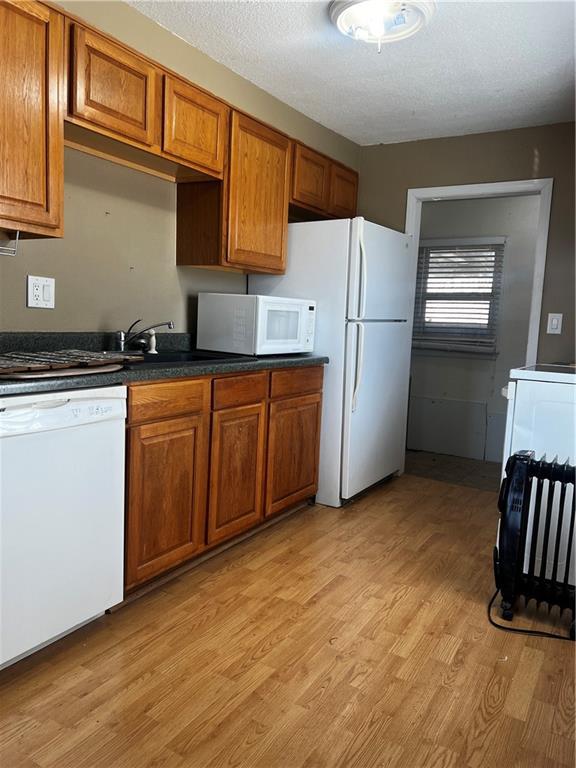 kitchen with a textured ceiling, dark countertops, white appliances, brown cabinetry, and light wood finished floors