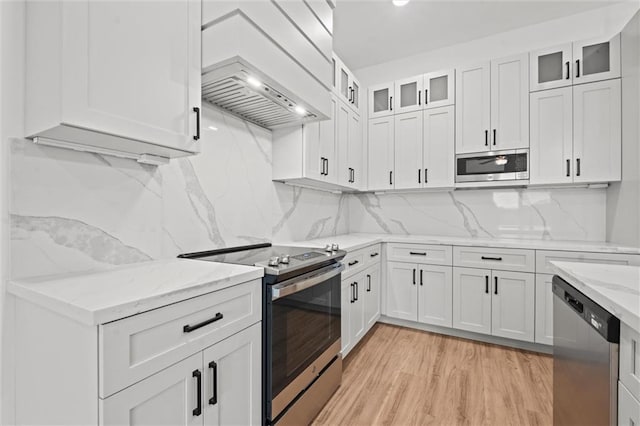 kitchen featuring premium range hood, light wood-type flooring, light stone counters, appliances with stainless steel finishes, and white cabinets