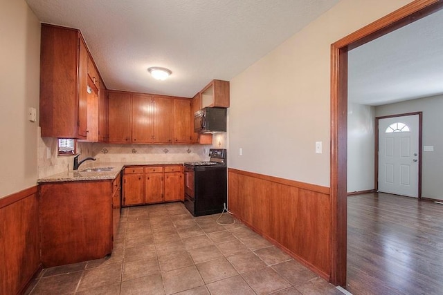 kitchen featuring black appliances, light stone counters, wooden walls, brown cabinetry, and wainscoting