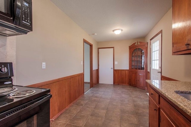 kitchen featuring a wainscoted wall, brown cabinets, black appliances, dark tile patterned floors, and wooden walls