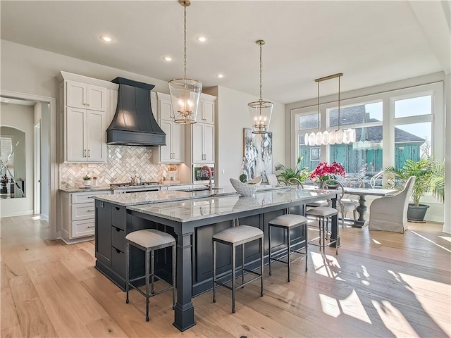 kitchen with custom exhaust hood, light wood-style flooring, a large island with sink, a sink, and tasteful backsplash