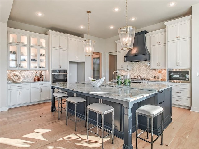 kitchen featuring premium range hood, a large island with sink, appliances with stainless steel finishes, light wood-style floors, and white cabinetry