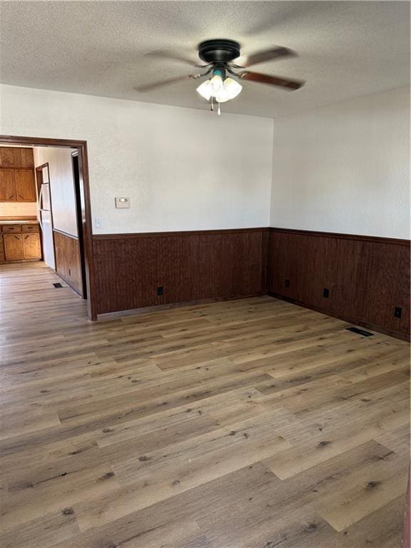 spare room featuring a textured ceiling, light wood-style flooring, a ceiling fan, and wainscoting