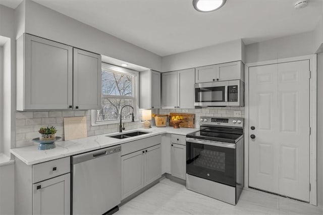 kitchen with gray cabinetry, a sink, tasteful backsplash, stainless steel appliances, and light stone countertops