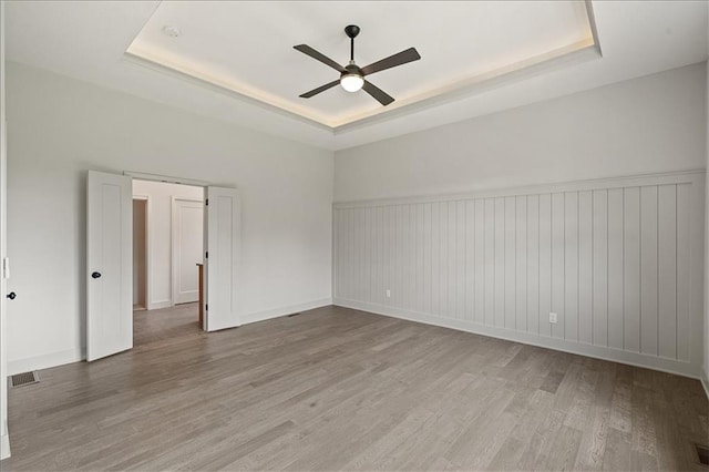 empty room featuring visible vents, a tray ceiling, wood finished floors, wainscoting, and ceiling fan