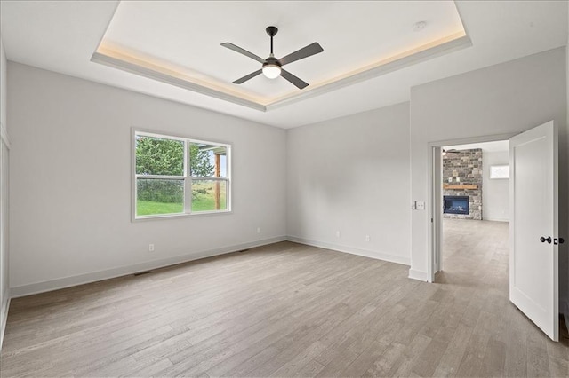 spare room featuring a raised ceiling, a ceiling fan, light wood-style floors, a stone fireplace, and baseboards