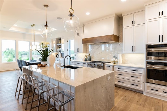 kitchen with custom range hood, light wood-style flooring, stainless steel appliances, a raised ceiling, and a sink