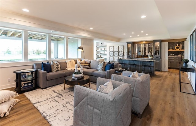 living room featuring baseboards, a tray ceiling, recessed lighting, indoor wet bar, and light wood-type flooring