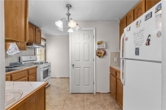 kitchen with white appliances, tile countertops, brown cabinetry, under cabinet range hood, and a chandelier