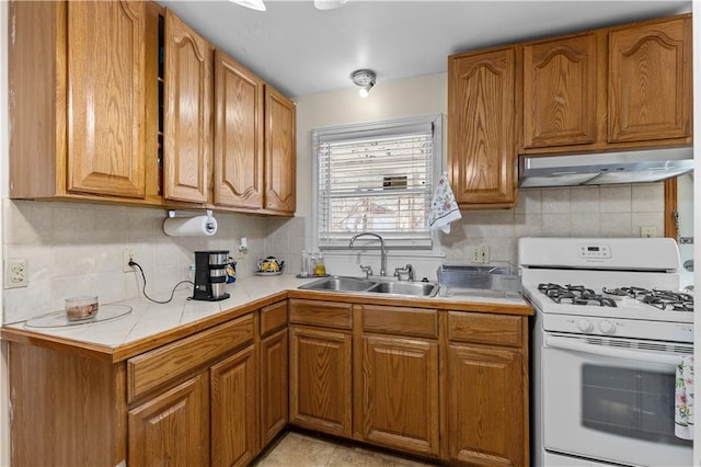 kitchen with brown cabinetry, white gas stove, a sink, under cabinet range hood, and tasteful backsplash