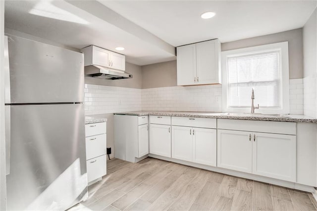 kitchen featuring freestanding refrigerator, a sink, under cabinet range hood, white cabinetry, and backsplash