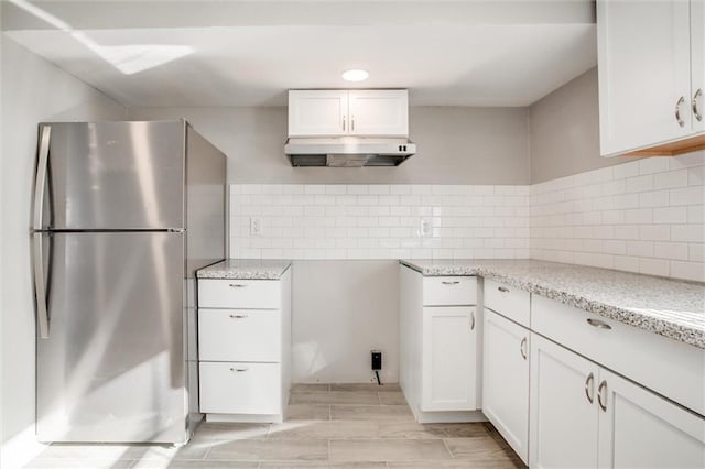 kitchen with under cabinet range hood, white cabinetry, freestanding refrigerator, decorative backsplash, and light stone countertops