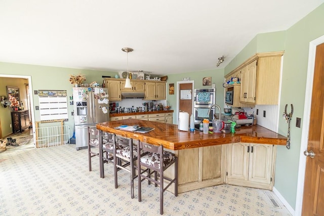 kitchen with dark countertops, under cabinet range hood, light brown cabinetry, a peninsula, and stainless steel fridge