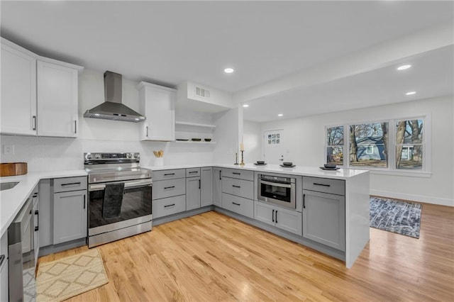 kitchen featuring gray cabinetry, a peninsula, stainless steel appliances, wall chimney exhaust hood, and open shelves