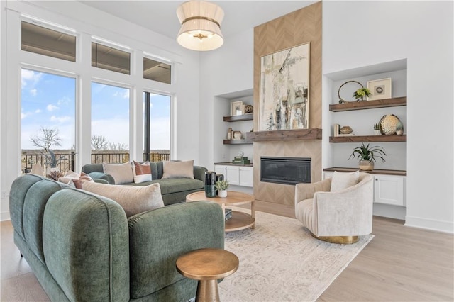 living room featuring built in features, baseboards, a high ceiling, light wood-style flooring, and a glass covered fireplace