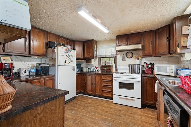 kitchen with a textured ceiling, dark countertops, white appliances, and under cabinet range hood