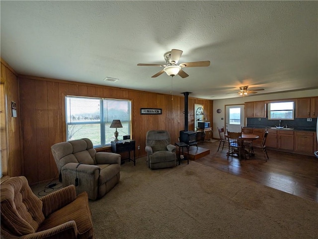 carpeted living area featuring visible vents, ceiling fan, wood walls, a wood stove, and a sink