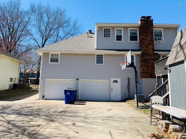 back of house featuring concrete driveway, an attached garage, a chimney, and a shingled roof