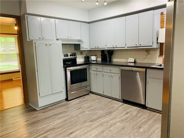 kitchen featuring dark countertops, under cabinet range hood, light wood finished floors, appliances with stainless steel finishes, and backsplash