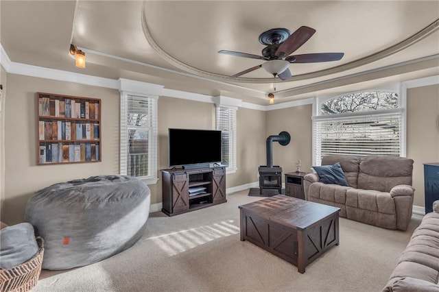 carpeted living area with crown molding, a wood stove, baseboards, a raised ceiling, and ceiling fan