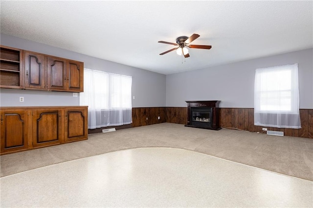 unfurnished living room featuring light colored carpet, ceiling fan, a wainscoted wall, and visible vents