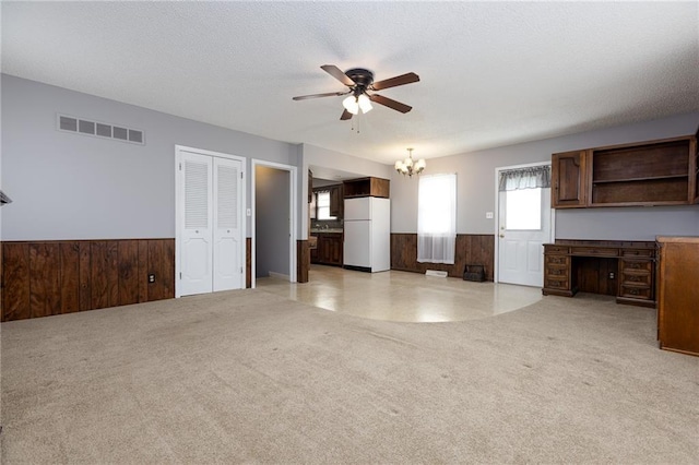 unfurnished living room featuring ceiling fan with notable chandelier, light carpet, a wainscoted wall, wood walls, and visible vents