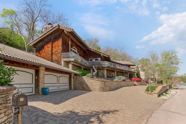 view of front of home with a chimney, a balcony, decorative driveway, an attached garage, and stone siding