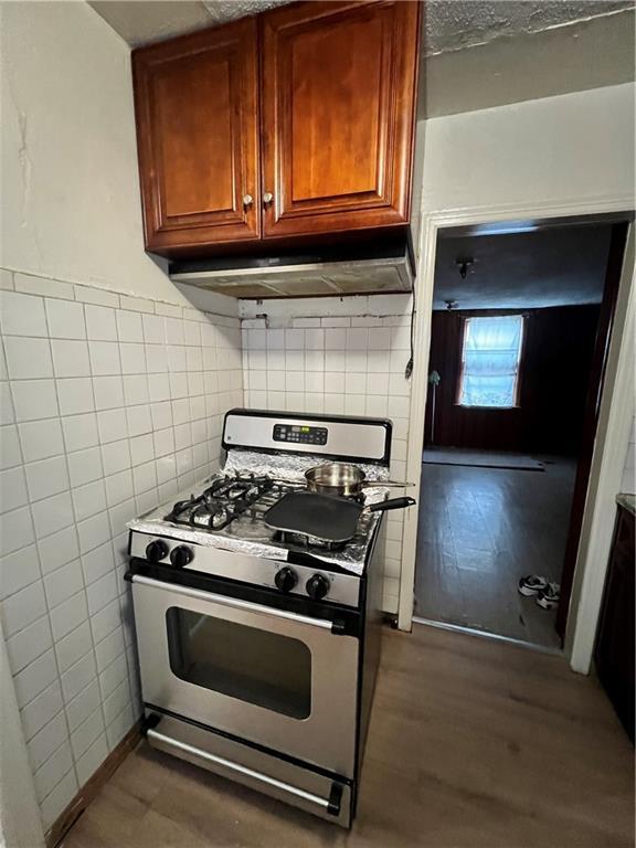 kitchen featuring tile walls, under cabinet range hood, brown cabinets, stainless steel gas stove, and dark wood-type flooring
