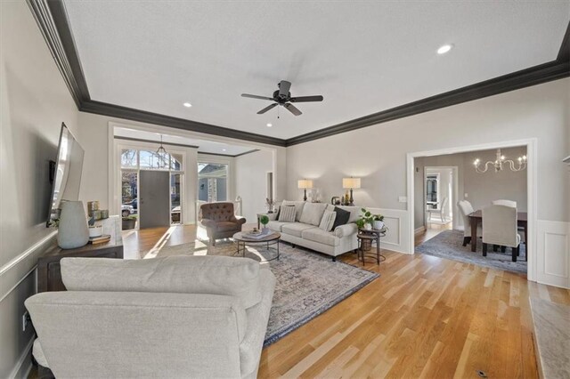 living room featuring crown molding, a wainscoted wall, light wood-style floors, and ceiling fan with notable chandelier