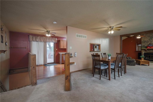 dining room with light colored carpet, a textured ceiling, a stone fireplace, a ceiling fan, and visible vents