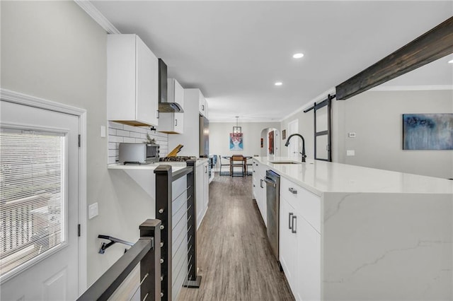 kitchen featuring a sink, crown molding, white cabinetry, a barn door, and wall chimney range hood