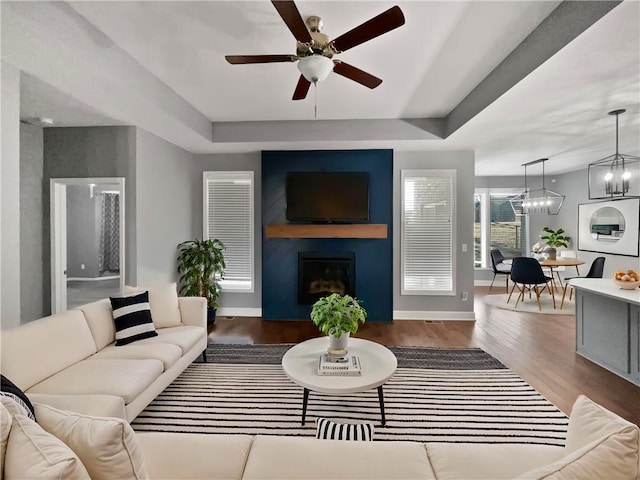 living area with ceiling fan with notable chandelier, a tray ceiling, a fireplace, and wood finished floors
